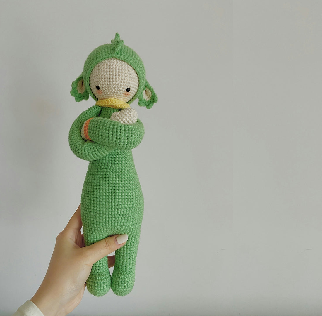 a hand holding a green stuffed animal in front of a white wall