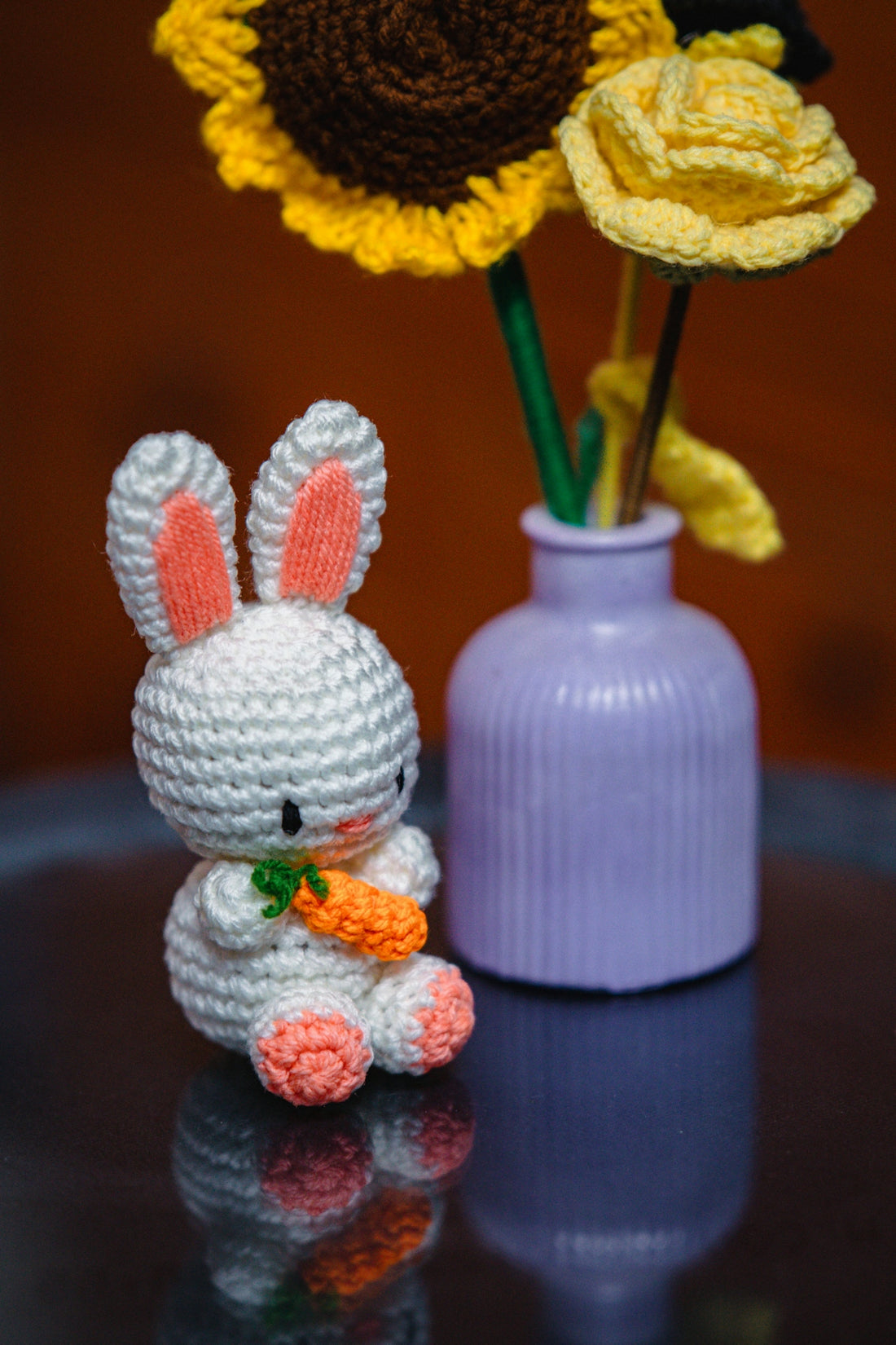 A crochet bunny holds a carrot near flowers.