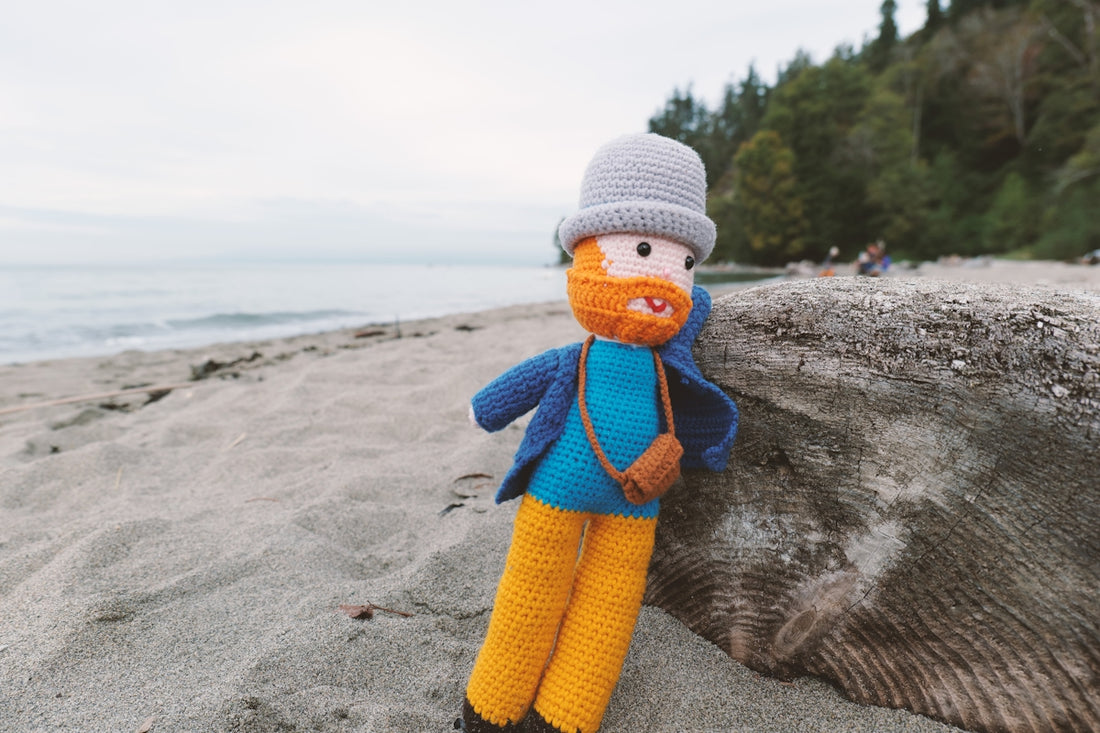 Crocheted doll with hat and camera on a sandy beach.
