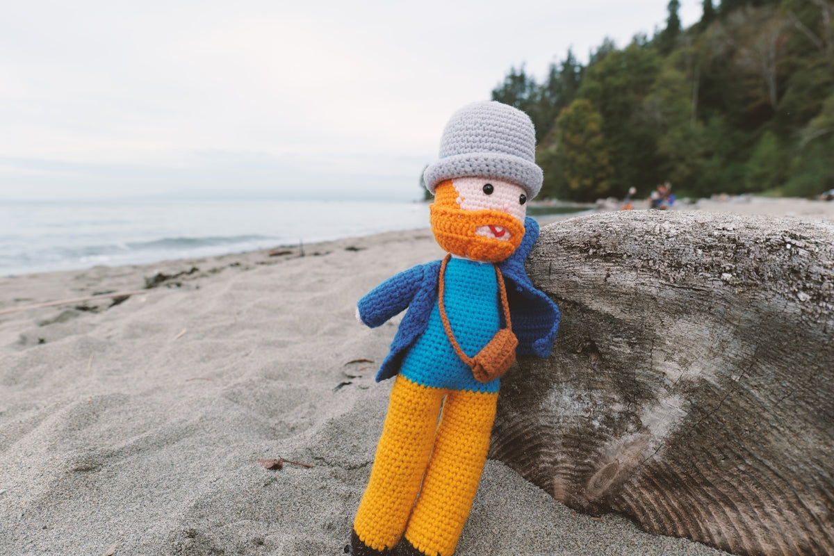 Crocheted doll with hat and camera on a sandy beach.